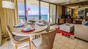 Un coin repas avec vue sur l’océan, un salon avec un canapé et une télévision, et un balcon avec des rideaux.