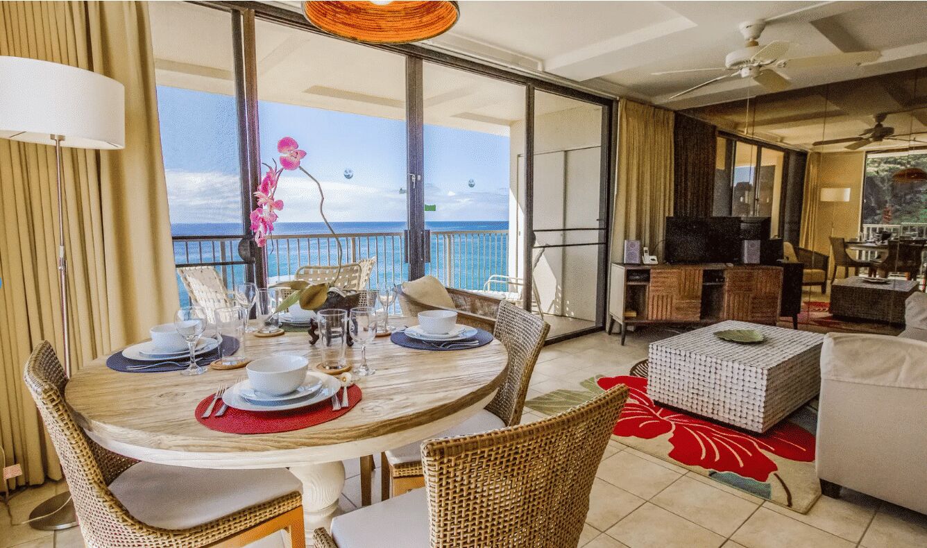 A dining area with a table set for four, a balcony with a view of the ocean, and a living area with a sofa and a television.