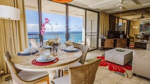 Un coin repas avec une table dressée pour quatre personnes, un balcon avec vue sur l’océan et un salon avec un canapé et une télévision.