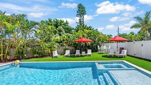 A swimming pool with clear blue water, surrounded by white lounge chairs and red umbrellas, set against a backdrop of lush greenery and a white fence.