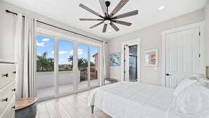 A bedroom with a large bed, sliding glass doors to a balcony, a ceiling fan, and a view of palm trees and buildings.