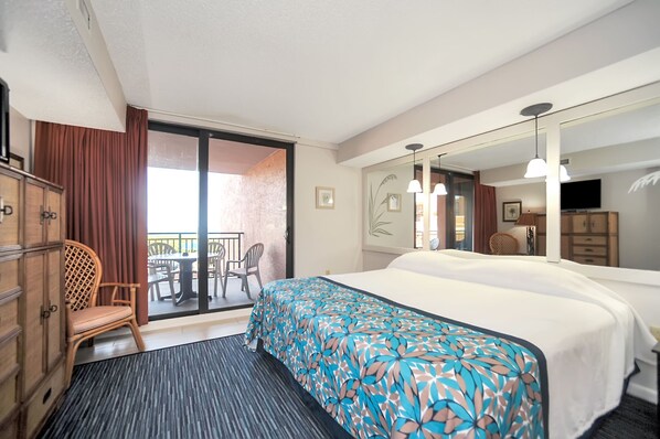 A hotel room with a large bed, a balcony with a table and chairs, a wardrobe, and a television. - Beachfront Condo w/ Pool & Balcony in FL (Indian Harbour Beach)