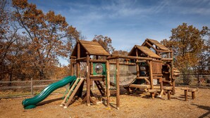 Una estructura de juegos de madera con un tobogán y elementos para escalar, rodeada de árboles y un cielo despejado.