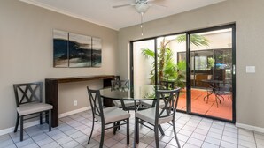 A dining area with a glass table and chairs, a ceiling fan, a painting on the wall, and a sliding glass door leading to an outdoor patio with plants.