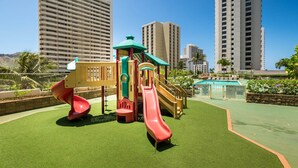 Un terrain de jeux avec des toboggans et des structures d’escalade, entouré de grands immeubles et d’une piscine.