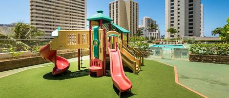 A playground with slides and climbing structures, surrounded by tall buildings and a pool.