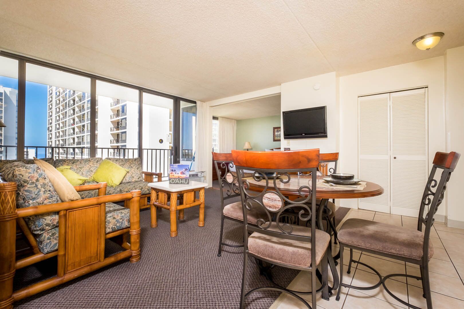 Et hotelværelse med balkon, spiseplads med rundt bord og stole, samt et fjernsyn monteret på væggen.