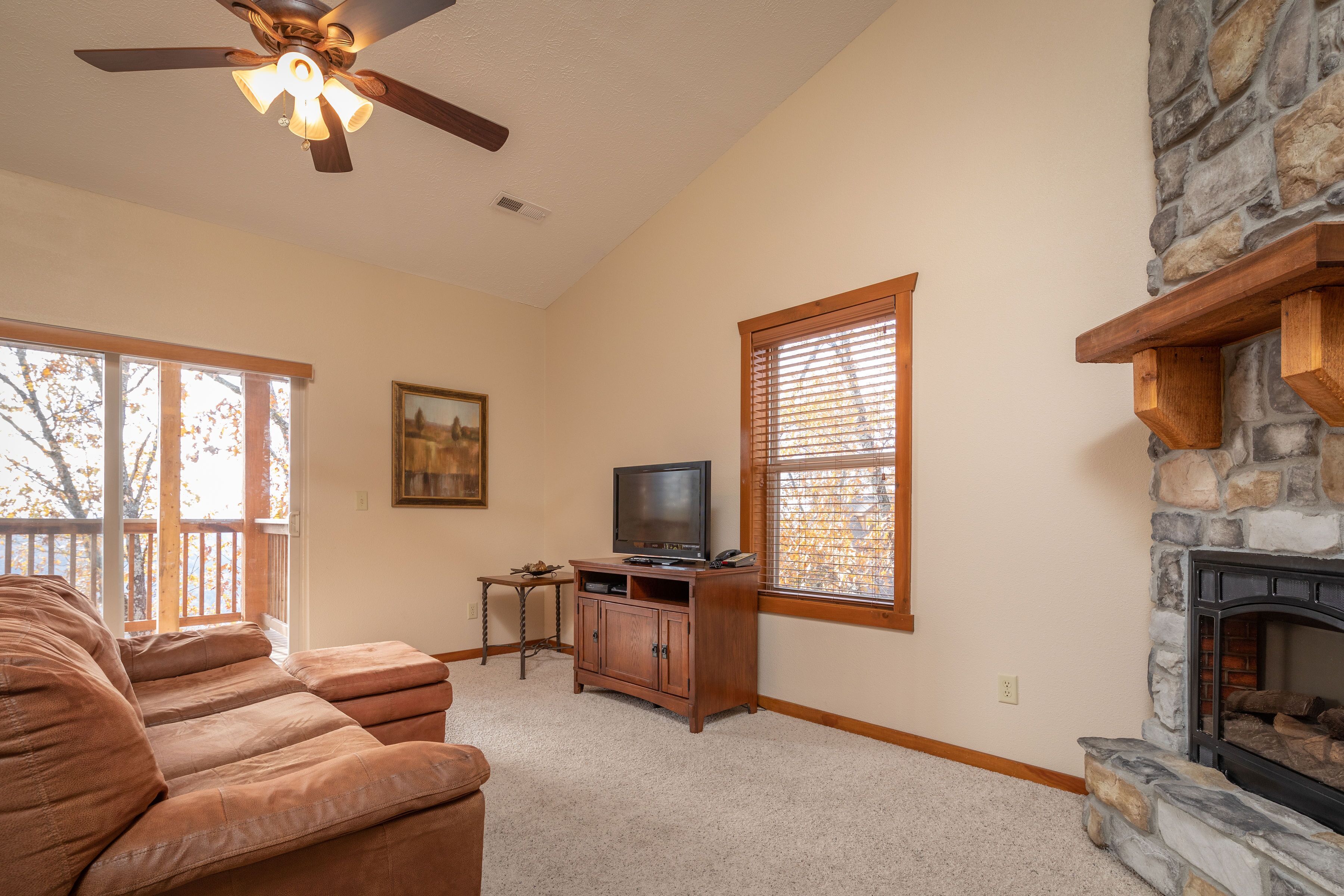 A living room with a stone fireplace, a television on a wooden stand, and a large window with blinds.