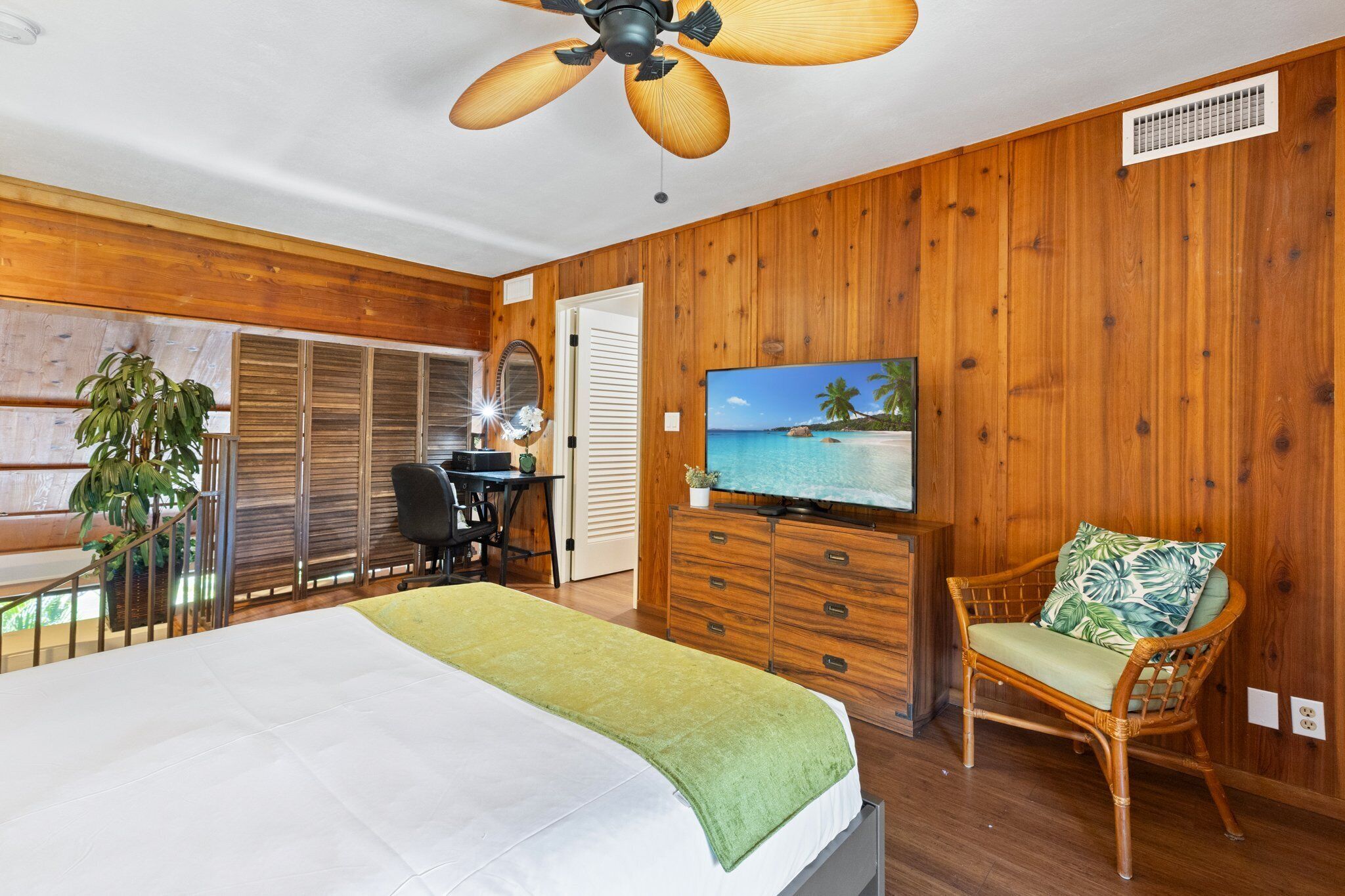 Un dormitorio con cama, televisor, pared de madera, ventilador de techo y ventana con persianas.