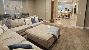 Un salon spacieux avec un grand canapé d’angle, un ventilateur de plafond et une vue sur la cuisine.