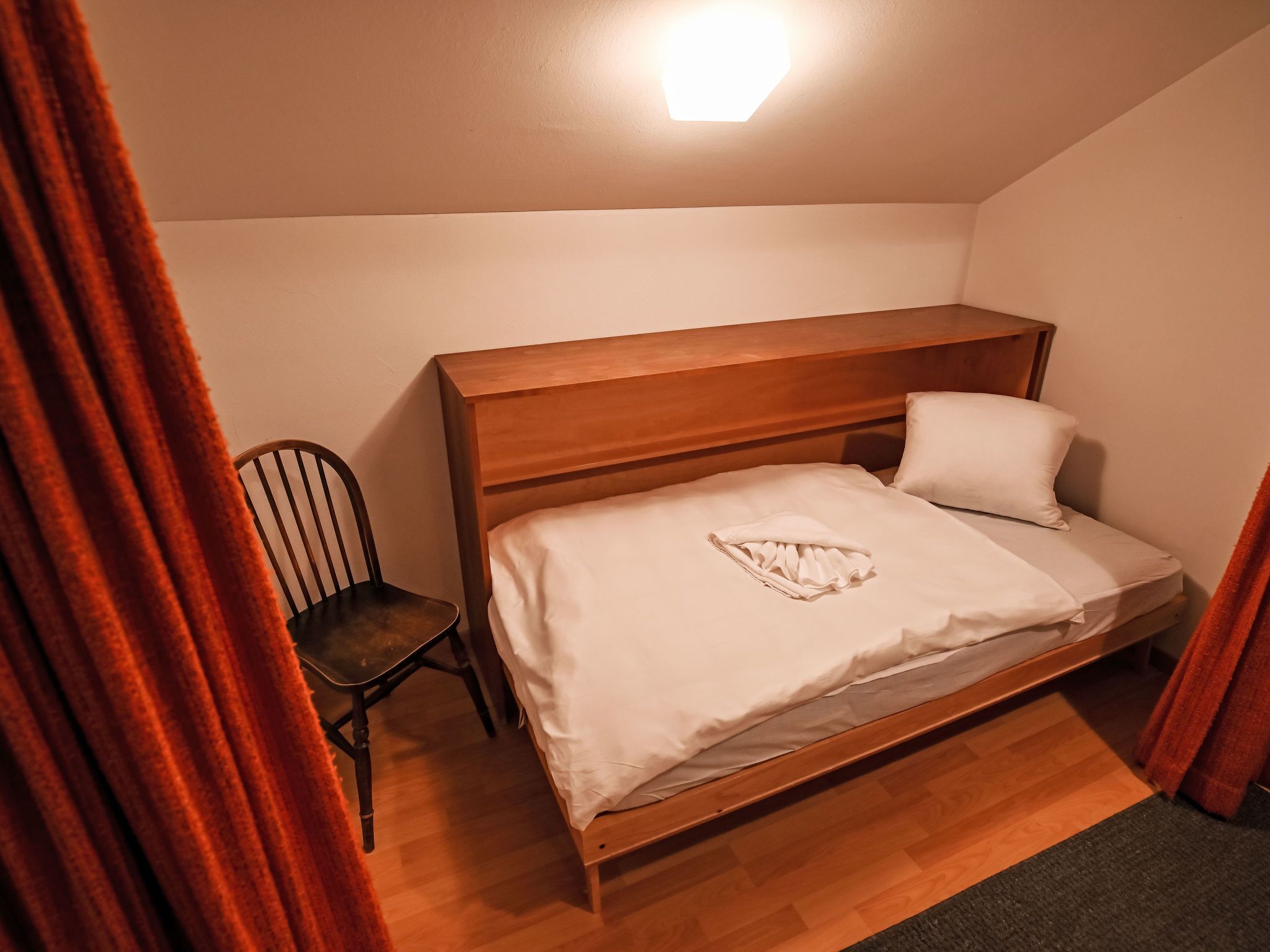 A small, single-bed room with a wooden headboard, a chair, and a bedside table.