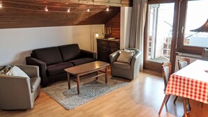 A cozy living room with a wooden ceiling, a sofa, a small table, a chair, and a dining table with a checkered tablecloth. - La Prada Wohnung 119 by Interhome (Laax)