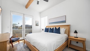 A bedroom with a large bed, a desk, and a chair. There is a window with blinds, a ceiling fan, and a painting on the wall.