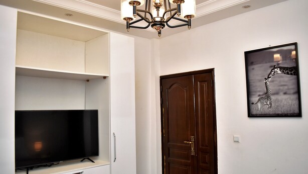 Una habitación con televisión de pantalla plana sobre un mueble blanco, una puerta de madera y un cuadro de jirafa en la pared.