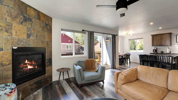 A modern living room with a fireplace, a sofa, a chair, and a dining area with a kitchen in the background. - New! Home located off Flathead River with balcony & fireplace (Columbia Falls)