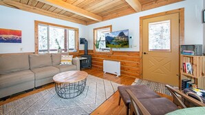 Ein gemütliches Blockhaus-Interieur mit Holzbalkendecke, einer Couch, einem runden Couchtisch, einem Kamin und einem Bücherregal.