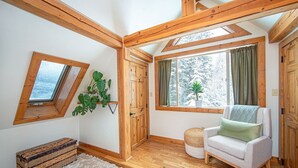 Ein gemütliches Zimmer mit Holzbalken, einem weißen, flauschigen Teppich, einer Korbsitzbank, einem weißen Sessel und Blick auf verschneite Bäume durch ein Fenster.