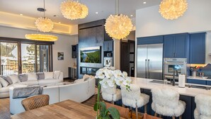 Ein modernes Wohnzimmer mit einem Holzesstisch, einem weißen Sofa und weichen Sesseln. Ein großer Fernseher und eine Küche mit blauen Schränken und Edelstahlgeräten sind ebenfalls vorhanden.