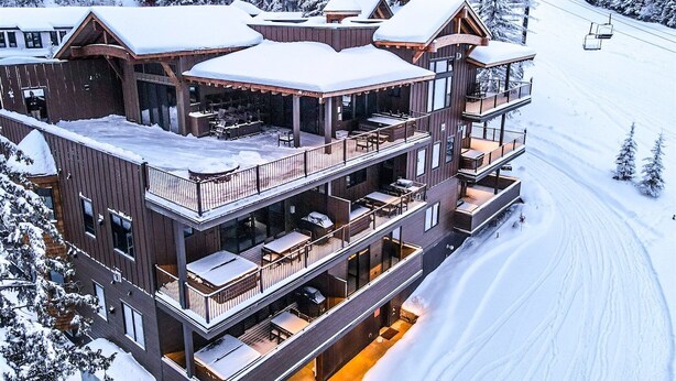 Ein mehrstöckiges Chalet mit Balkonen, ein Sessellift im Hintergrund und eine verschneite Waldlandschaft.