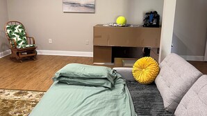 Un dormitorio con una cama, un sofá gris, una mecedora de madera, un cojín amarillo y un cuadro en la pared.
