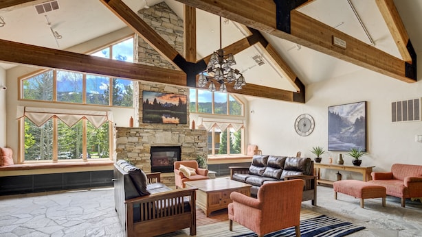 A spacious living room with stone flooring, wooden beams, large windows, a fireplace, and comfortable seating. - Gateway Studio | Pool, Hot Tub, Walk to Slopes! (Keystone)