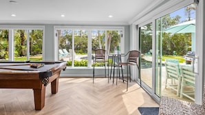 A pool table in a room with large windows overlooking a pool and garden.