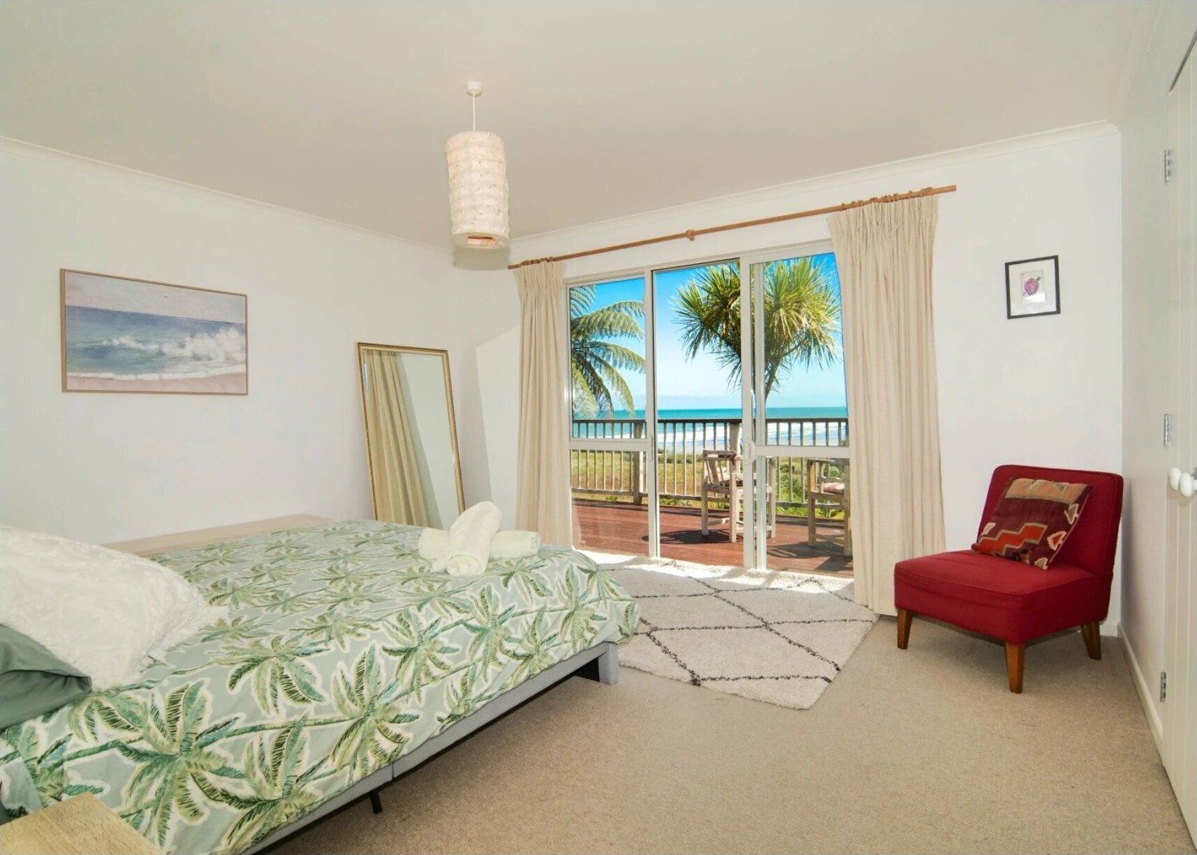 A bedroom with a bed, a red chair, a large window with a view of palm trees and the sea, and a ceiling light.