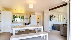 A modern kitchen with white cabinetry, a dining area with a white table and bench, and a living area with a sofa and a guitar.