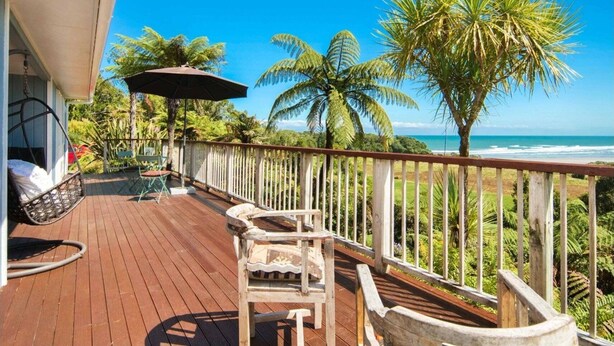 A wooden deck with a hanging chair and a table, overlooking a beach with palm trees and a clear blue sky.