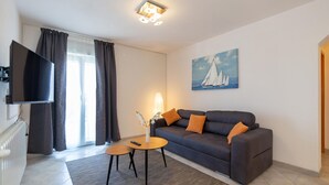 Ett modernt vardagsrum med en mörkgrå soffa, två orange kuddar, ett litet träbord och en golvlampa. En tavla med segelbåtar hänger på väggen, och en platt-TV är monterad på väggen.