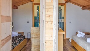 Ein Schlafzimmer einer Holzhütte mit zwei Betten, einer zentralen Holzsäule und einem durch eine Glastür sichtbaren Badezimmer.