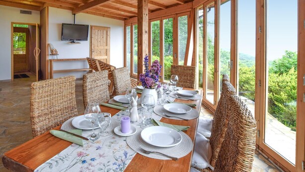 Ein Essbereich mit einem Holztisch, gedeckt für vier Personen, umgeben von Korbstühlen, mit Blick auf Grün durch große Fenster.