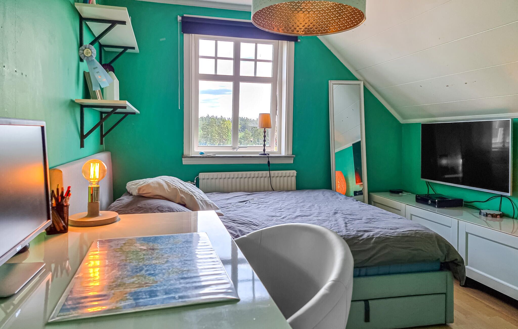 Ein Zimmer mit Bett, Schreibtisch, Stuhl und Fernseher. Ein Fenster bietet Blick auf Bäume, auf dem Schreibtisch steht eine Lampe.