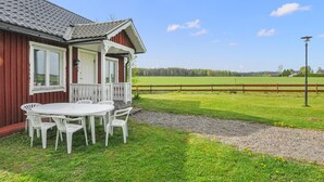 Uma casa com varanda, uma mesa com cadeiras e vista para um campo verde e árvores.