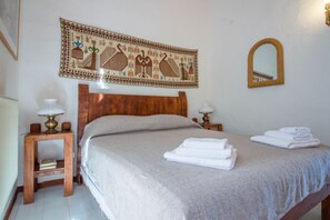 Een netjes opgemaakt bed met hoofdbord, twee nachtkastjes met lampen, een wandtapijt, een spiegel en opgevouwen handdoeken.