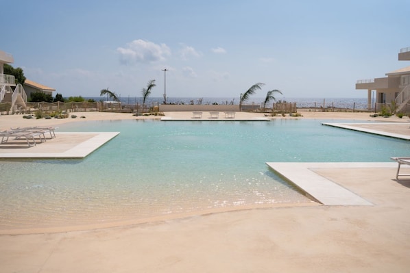 Una piscina con vistas despejadas al mar, rodeada por una zona de arena con sillas de descanso y un edificio con balcón.