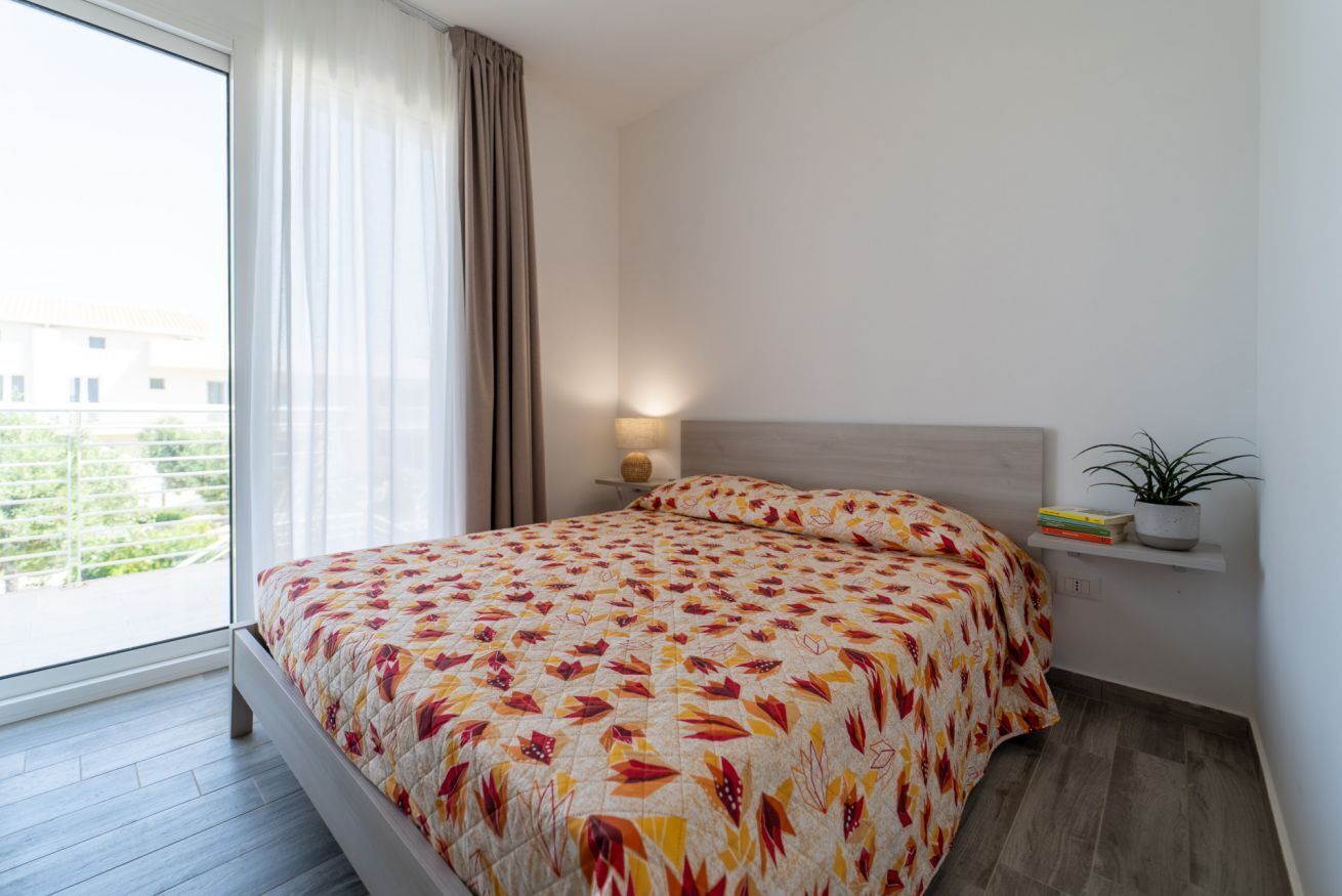 A bedroom with a bed, a nightstand, a lamp, a window with curtains, and a view of buildings outside.