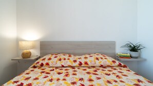 Ein Bett mit gemustertem Überwurf, eine Nachttischlampe, eine kleine Ablage mit Büchern und einer Pflanze sowie ein Holz-Kopfbrett.