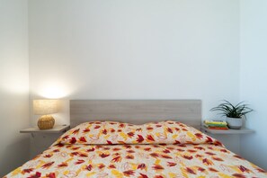 Een bed met een geborduurd sprei, een bedlampje, een klein plankje met boeken en een plant, en een houten hoofdbord.