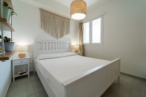 Una cama blanca con cabecera, una lámpara de noche, una mesita de noche, un colgador de macramé en la pared y una ventana con cortinas.