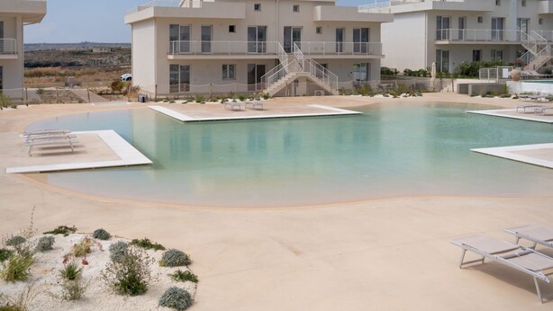 A pool area with a clear sky, a building with multiple balconies, and a staircase leading to the upper floor. - GuestHost - Cozy 40 sqm seaview flat, able to accommodate up to 3 people, located inside the renowned Borgo 84 residence, equipped with swimming pool and cycle path. The property is located just 3 km from the center of Marzamemi, one of the most fascinat (Pachino)