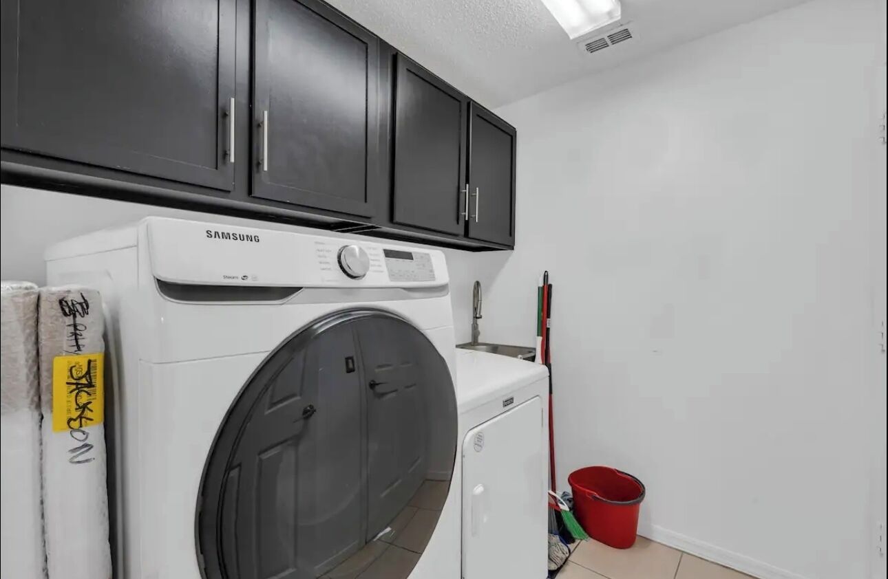 Een wasserette met een Samsung wasmachine, donkere kastjes, een rode emmer en een witte muur.