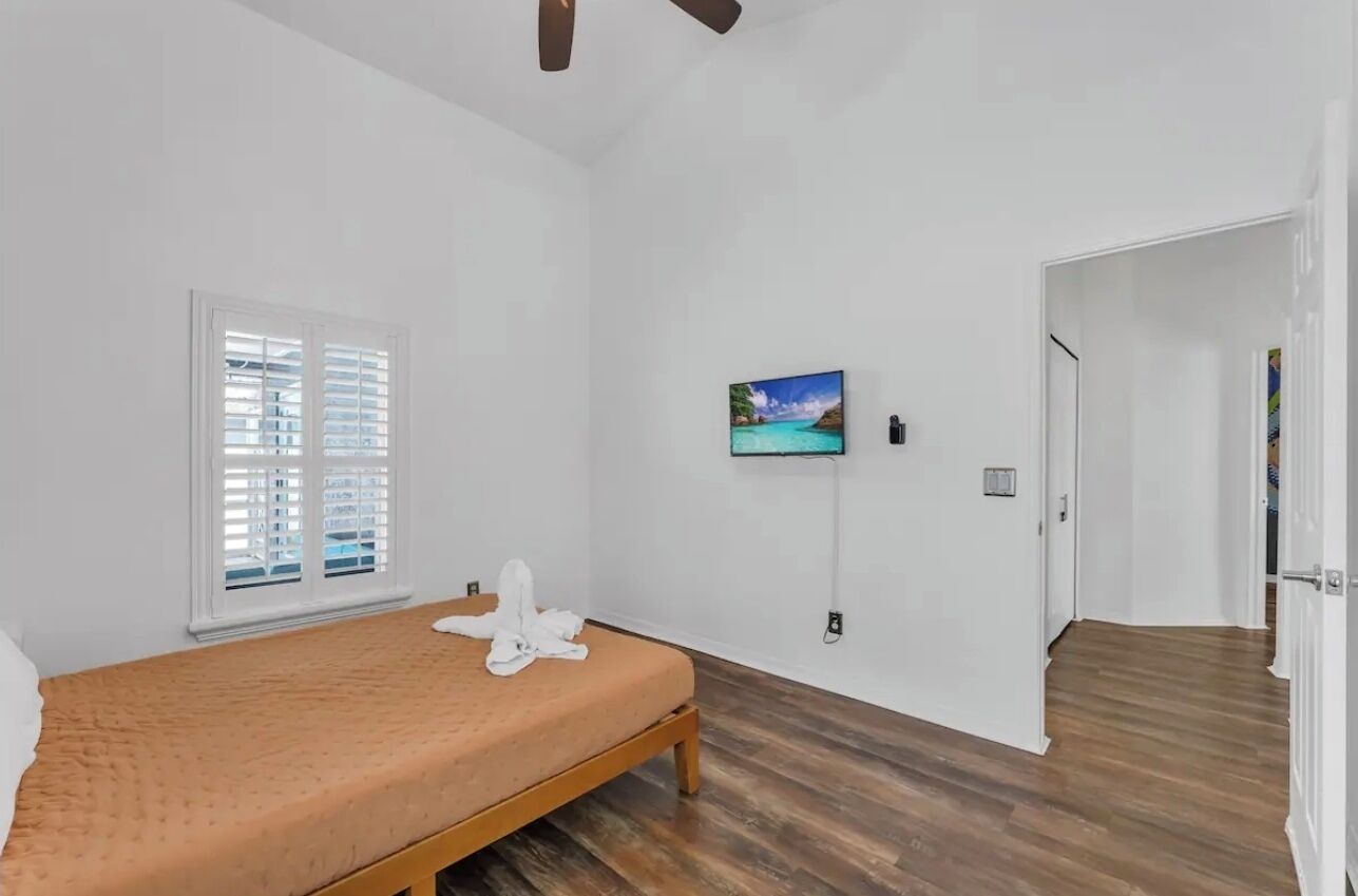 Ein Schlafzimmer mit Bett, einem Fenster mit Jalousien, einem an der Wand befestigten Fernseher und Holzboden.