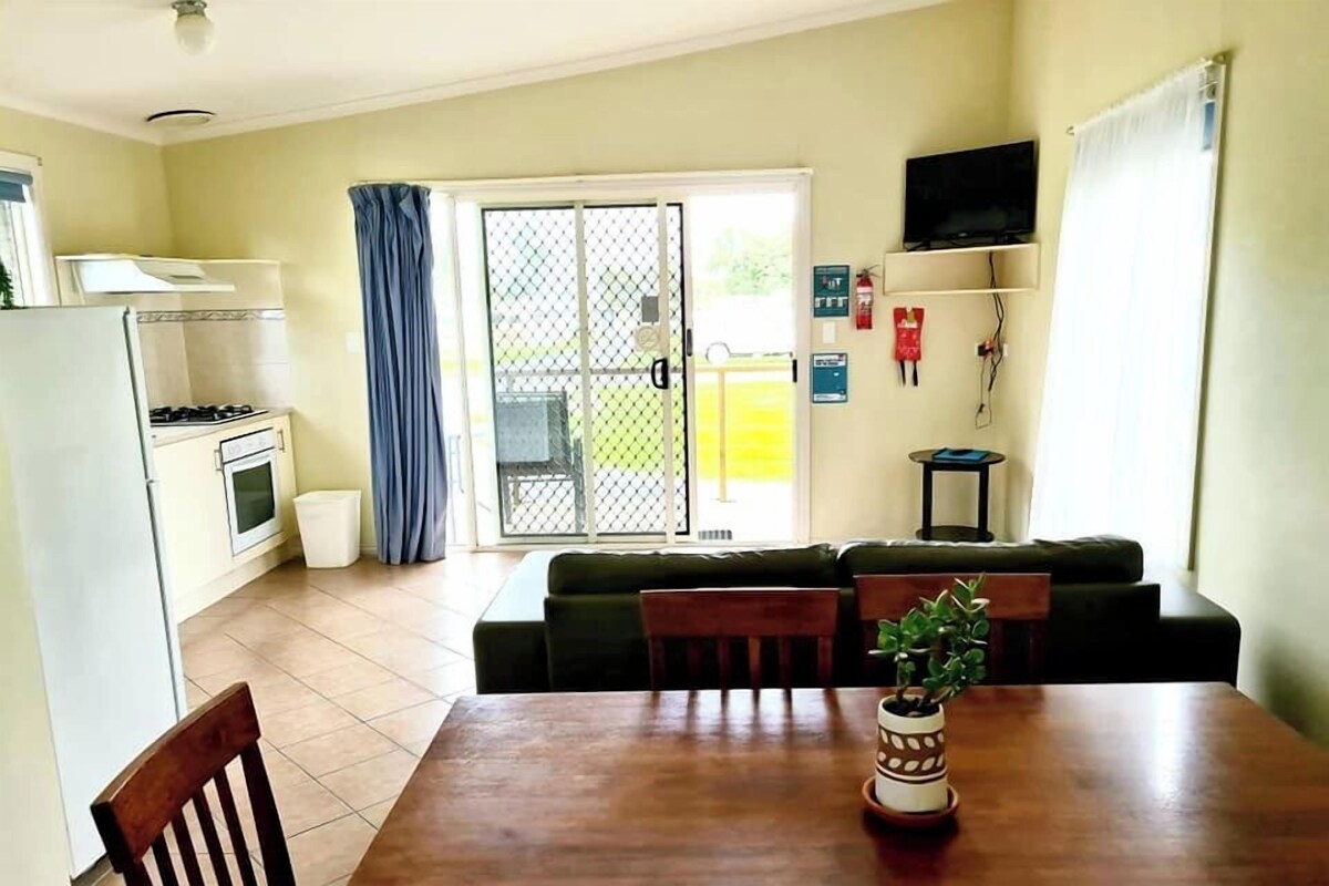 A compact kitchen with a refrigerator, stove, and oven, a dining table with chairs, a sofa, and a TV on the wall.