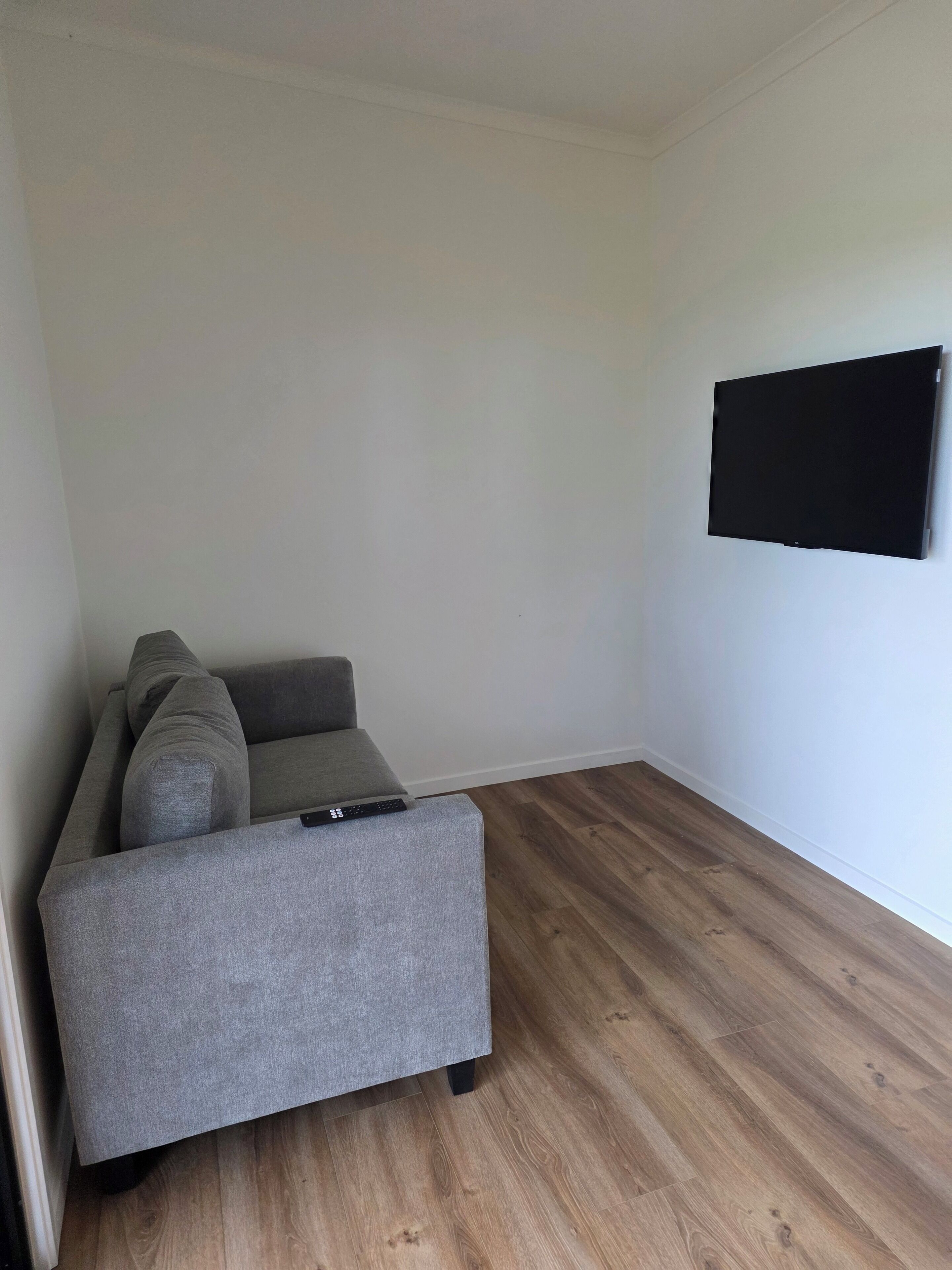 Een kamer met een grijze bank, een flatscreen televisie aan de muur en houten vloeren.