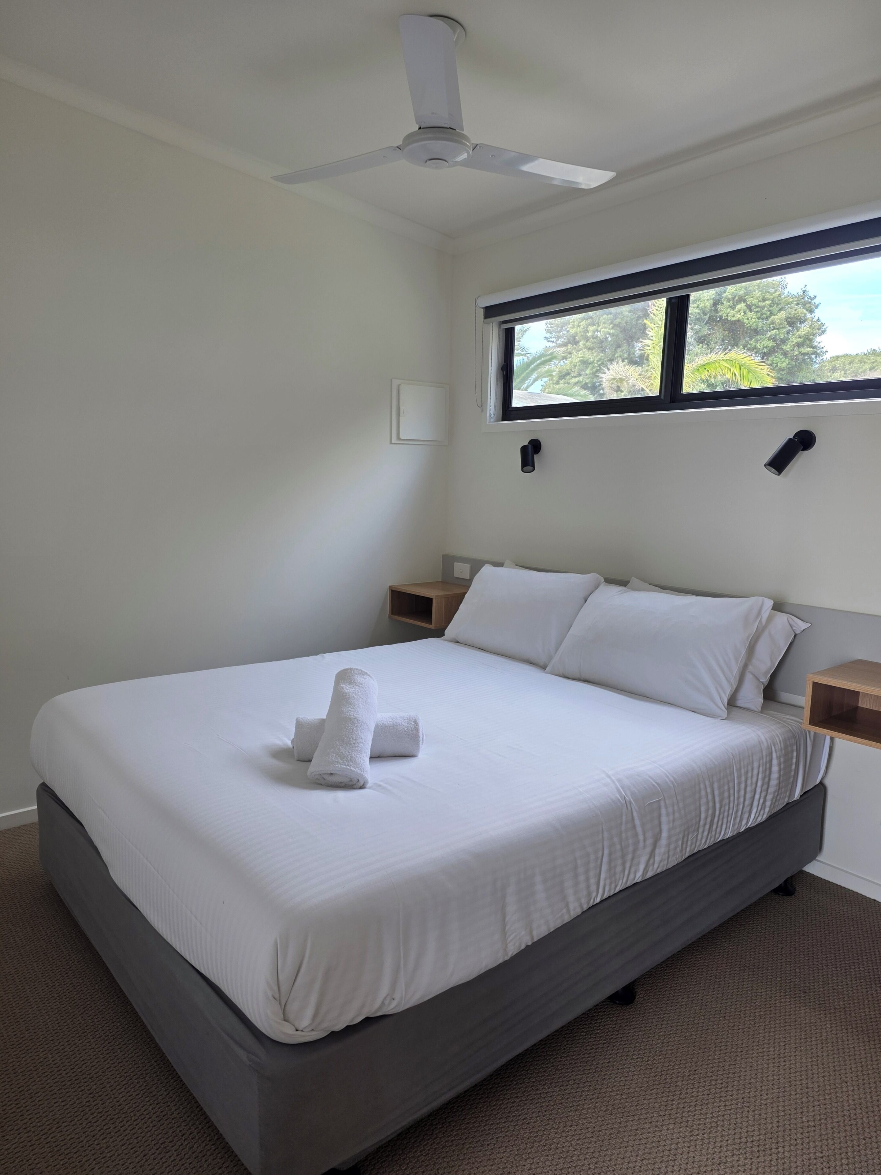 A bedroom with a bed, a ceiling fan, a window, and a nightstand.