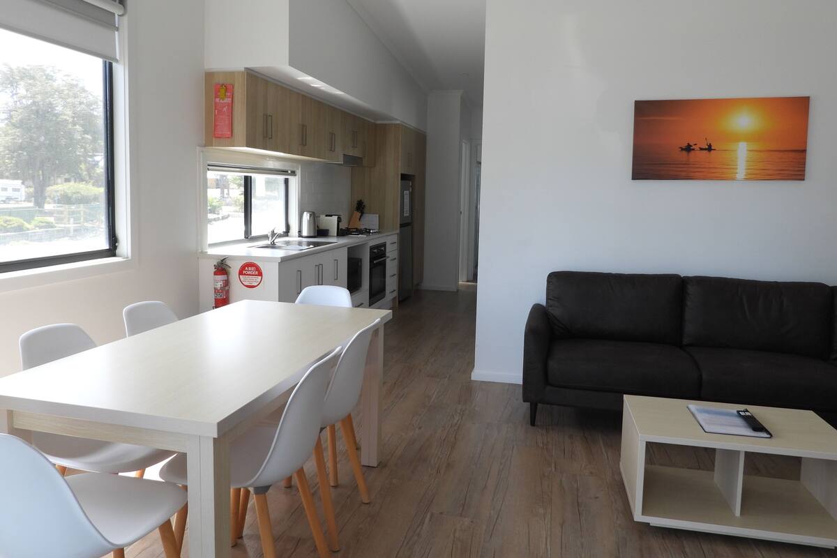 Een moderne keuken met witte kasten, een eethoek met een witte tafel en stoelen, een zwarte bank en een schilderij van een zonsondergang.