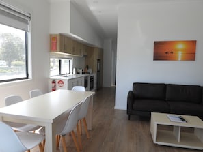 Una cocina moderna con armarios blancos, un comedor con mesa y sillas blancas, un sofá negro y un cuadro de una puesta de sol.