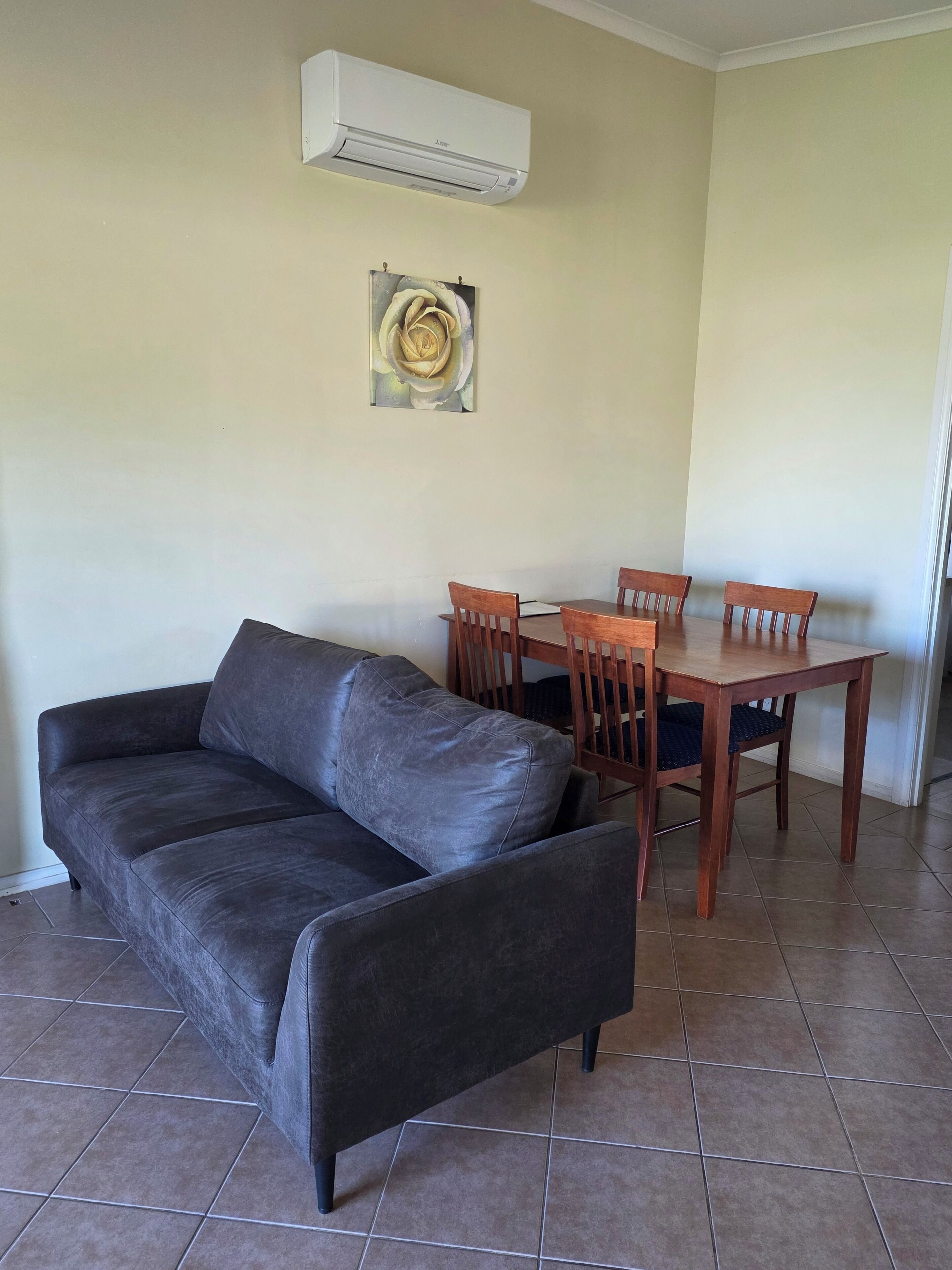 A living room with a dark leather sofa, a wooden dining table with four chairs, and an air conditioning unit mounted on the wall.