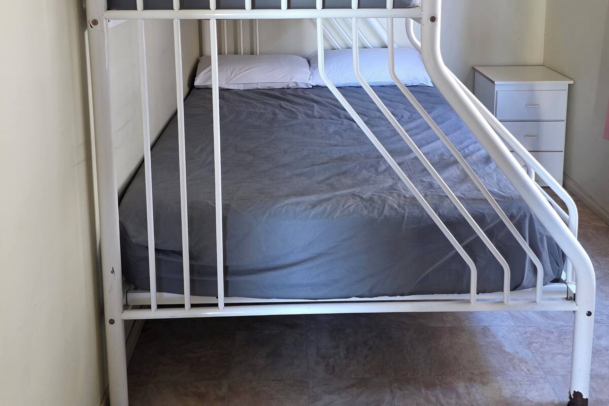 Een wit metalen bed met grijs beddengoed, een klein nachtkastje en een ladekast.